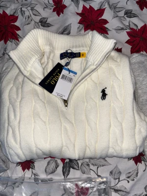Vit kabelstickad tröja Polo Ralph Lauren - Kabelstickad vit tröja från Polo Ralph Lauren med halv dragkedja och hög krage. Klassisk svart logga broderad på bröstet. Tröjan är långärmad och har ribbade muddar och nederkant. Perfekt för dig som gillar stilren och tidlös design.