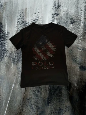 Svart Polo t-shirt - Svart t-shirt från Polo med coolt tryck av en örn i rött, vitt och blått på bröstet. Klassisk rund hals och korta ärmar. Perfekt för dig som vill sticka ut med en unik graphic tee.