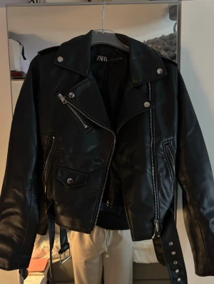 Svart skinnjacka från Zara - En svart skinnjacka från Zara. Jackan har silvriga detaljer, flera dragkedjor, knappar och ett bälte nedtill. Långärmad modell.
