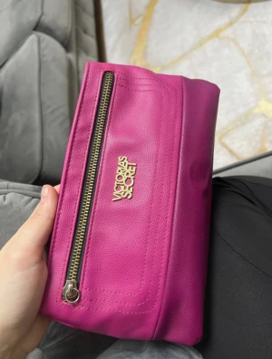 Rosa kuvertväska från Victoria's Secret - Snygg rosa kuvertväska från Victoria's Secret i skinnimitation med guldfärgad logga och dragkedja framtill. Väskan har magnetlås och ett rymligt fack, perfekt för det viktigaste. Stilren och trendig design som sticker ut.