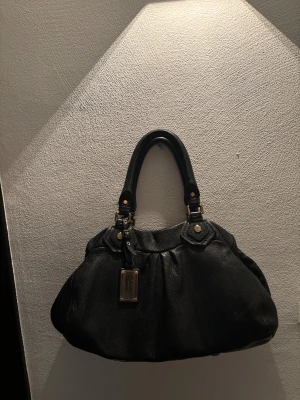 Marc Jacobs väska  - Snygg svart handväska från Marc Jacobs i mjukt skinn med guldfärgade detaljer. Följer tyvärr inte med något band. 