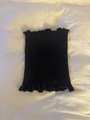 Söt tube topp/linne - Säljer en svart tubtopp med smockad struktur och volangkant upptill och nedtill. Du kan bära den både som bandeau eller med band. 