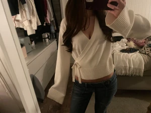 Vit omlott kofta med knyt - Säljer en vit omlott kofta med knyt i midjan och breda ribbade muddar. Koftan har v-ringad hals och är croppad i modellen, vilket ger en snygg och modern look. Materialet är stickat och mjukt, perfekt för att hålla dig varm och stilren. Passar S/M men är i storlek 170!