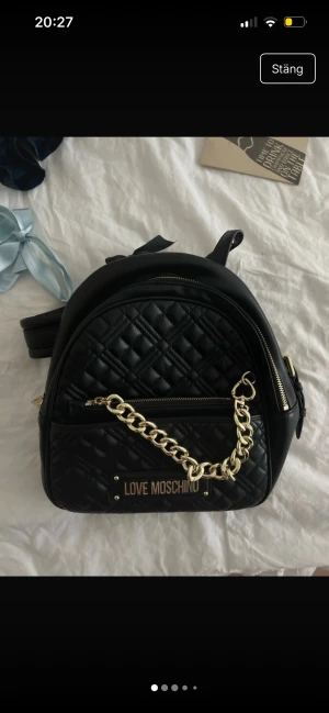 Svart quiltad ryggsäck Love Moschino - Stilren svart quiltad ryggsäck från Love Moschino i skinnimitation med guldfärgade detaljer och kedja framtill. Väskan har dragkedja, justerbara axelband och ett innerfack med snygg logodetalj. Perfekt för dig som vill ha en trendig och praktisk accessoar.