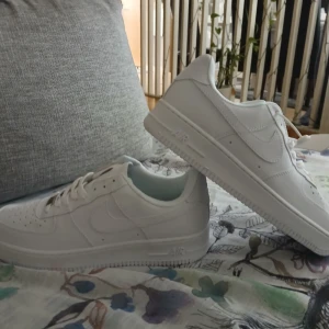 Nike Air Force 1 vita sneakers - Nike Air Force 1 sneakers i helvitt med klassisk låg siluett. Skorna har snörning, rund tå och platt sula med det ikoniska Air-märket på sidan. Tillverkade i skinn med detaljer i syntet och textil på insidan. Perfekt för dig som gillar clean och stilren streetwear.