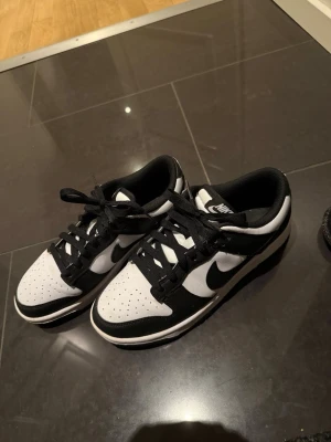 Nike Dunk Low svart/vit sneakers - Nike Dunk Low sneakers i svart och vitt med klassisk låg siluett. Skorna har snörning, perforerad tå och en platt sula. Swoosh-loggan syns tydligt på sidan och materialet är en mix av syntet och skinn. Perfekt för dig som gillar streetwear och vill ha en clean look.