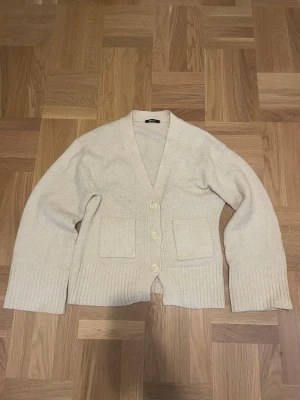 Beige stickad cardigan från Gina Tricot - Mysig beige stickad cardigan från Gina Tricot med v-ringad hals, stora ribbade muddar och tre knappar framtill. Två fickor på magen.