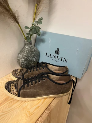 Lanvin sneakers med mocka och reptilmönster - Lanvin sneakers i brun mocka med svart reptilmönstrad tå och svarta snören. Snygga kontraster med beige sula och mörka detaljer. Klassisk rund tå och platt sula, perfekt för dig som gillar exklusiv streetstil med unika materialval. lite rivna där bak, ställ frågor‼️💬