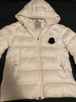 Vit Moncler pufferjacka M/S  - Säljer en snygg vit pufferjacka från Moncler med huva och dragkedja framtill. Jackan har två fickor med knappstängning och ett svart Moncler-märke på bröstet. Perfekt för kalla dagar och riktigt stilren look. Pris kan diskuteras och storleken är för personer med S/M