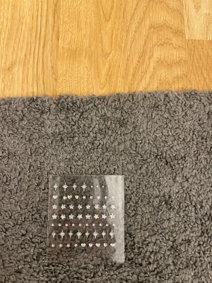 Små holografiska ansiktsdekaler - Ett ark med holografiska ansiktsdekaler i olika former som stjärnor, hjärtan och blommor. Perfekt för att skapa kreativa och glittriga sminklooks. Dekalerna är små och har en skimrande effekt som reflekterar ljus i regnbågens färger. Materialet är plast med självhäftande baksida.