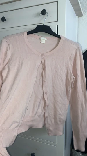 Ljusrosa kofta från H&M - Söt ljusrosa kofta från H&M med rund halsringning och knappar framtill. Koftan har långa ärmar och ribbade muddar vid ärmslut och nederkant. Perfekt att slänga över en topp för en chill look.