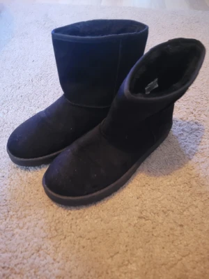 Svarta mockaboots med platt sula - Svarta boots i mocka med platt sula och rund tå. Skorna har en enkel och stilren design med mjukt foder på insidan, perfekta för kalla dagar. Yttersulan är grovt mönstrad för extra grepp. Passar dig som gillar en clean och mysig look. 