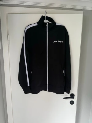 Palm Angels svart tracksuit set - Säljer ett svart tracksuit set från Palm Angels med vita ränder längs ärmar och ben. Jackan har hög krage, hel dragkedja och logga på bröstet. Byxorna har resår i midjan, logga på låret och dragkedjor vid bensluten. Snygg och sportig look med klassiska detaljer.