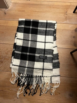 Rutig svartvit halsduk från Burberrys - Säljer en klassisk halsduk från Burberrys i svartvitt rutigt mönster. Tillverkad i 100% lammull och har fransar i ändarna. Perfekt accessoar för att lyfta din outfit med en tidlös brittisk vibe.