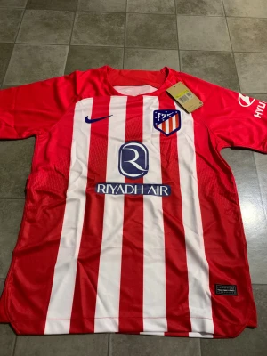 Rödvit Atlético Madrid fotbollströja Nike - Säljer en officiell Atlético Madrid matchtröja från Nike i storlek S. Tröjan har röda och vita vertikala ränder, klubbmärke på bröstet och flera sponsorer tryckta fram och bak. Materialet är lätt polyester, perfekt för fotboll och träning.