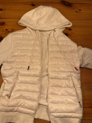 Vit Moncler Cardigan  - Säljer en snygg vit cardigan från Moncler med huva och dragkedja. Cardigan har quiltad framsida, två fickor med dragkedja och en Moncler-logga på ärmen. NFC tagen funkar. Inget fel på den. Oanvänd med. 
