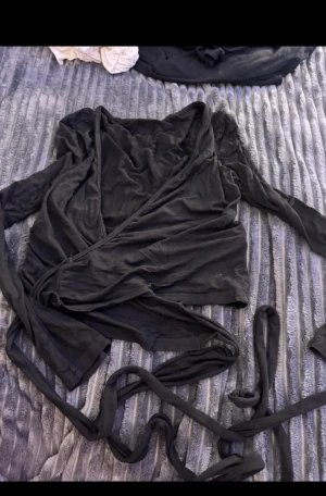 Svart omlottopp med långa band - Säljer en svart omlottopp med långa ärmar och extra långa knytband som kan snurras runt midjan för en snygg look. Toppen har en djup v-ringning och är gjord i mjukt, tunt material som känns skönt mot huden. Perfekt att styla med jeans eller kjol.