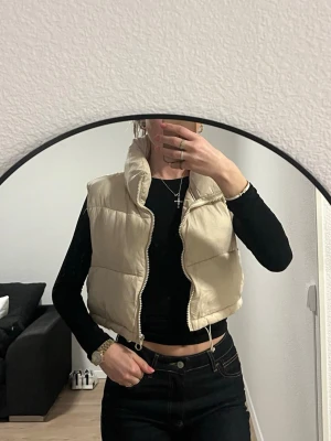 Beige dunväst med dragkedja - Snygg beige dunväst från Padded Jacket med hög krage och quiltad design. Västen har en croppad passform och stängs med dragkedja framtill. Perfekt att slänga över en hoodie eller långärmad tröja för en chill look.