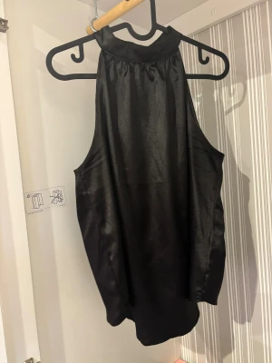 Svart halterneck blus i satin - Säljer en svart halterneck blus i glansigt satinmaterial. Toppen har en hög krage med knappstängning i nacken och är ärmlös med en snygg, flowy passform. Perfekt för dig som gillar stilrena och eleganta plagg med lite edge.
