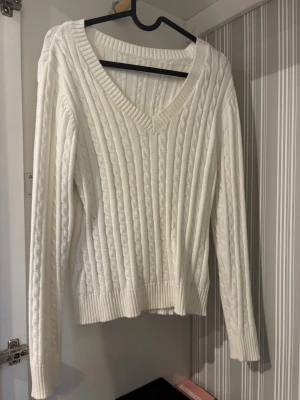 Vit kabelstickad v-ringad tröja - Kabelstickad vit tröja med v-ringning och långa ärmar. Tröjan har ribbade muddar vid ärmslut och nederkant, vilket ger en snygg och klassisk look. Perfekt för dig som gillar stilrena och mysiga plagg med lite preppy känsla.