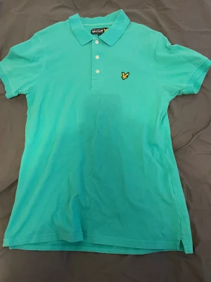 Turkos piké från Lyle & Scott - Säljer en fräsch turkos pikétröja från Lyle & Scott med klassisk krage och tre knappar framtill. Tröjan har korta ärmar och den ikoniska gula fågel-loggan broderad på bröstet. Perfekt för dig som gillar stilren och sportig look.