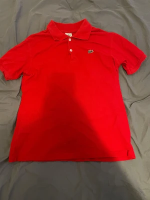 Röd piké från Lacoste - Säljer en klassisk röd pikétröja från Lacoste med den ikoniska krokodilloggan på bröstet. Tröjan har korta ärmar, krage och två knappar framtill. Perfekt för dig som gillar stilrena och sportiga plagg med en touch av street.