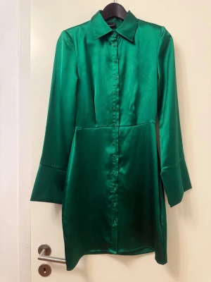 Grön skjortklänning i satin - Snygg grön skjortklänning från NA-KD i glansigt satinmaterial. Klänningen har lång ärm med bred manschett, klassisk krage och knappar hela vägen framtill. Passformen är normal och längden är kort, perfekt för dig som gillar stilrena och trendiga plagg.