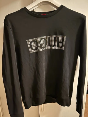 Svart tröja med HUGO-paljetter - Svart långärmad tröja från HUGO med coolt paljettmönster på bröstet som stavar HUGO. Tröjan har rund hals och är gjord i mjuk bomull, perfekt för dig som gillar stilrena och trendiga plagg med lite bling.