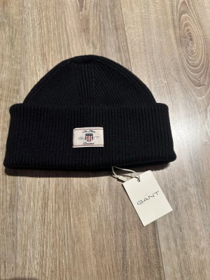 Svart ribbstickad mössa från GANT - Svart ribbstickad mössa från GANT med uppvikt kant och snyggt broderad patch framtill med logga och texten 'New Haven Connecticut'. Klassisk och stilren design som passar perfekt till kalla dagar.