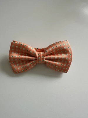 Orange fluga med blommigt mönster - Festlig orange fluga med mönstervävda små grönvita blommor / prickar.  Justerbart band för bästa passform. Ny i förpackning.   Hämtas i Stockholm eller skickas mot porto.