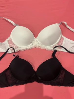 Svart och vit spetsdetaljerad bh-duo - Två snygga bh:ar, en i svart och en i vit, båda med spetsdetaljer längs kanten och sidorna. De har vadderade kupor och justerbara axelband för bästa passform. Perfekt för dig som gillar stilrena underkläder med en touch av spets. 