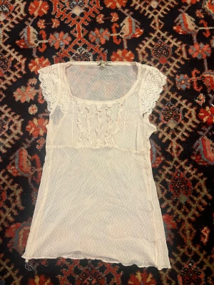 Ljusrosa spetsblus med volang från KRLINDE - Säljer en söt vit blus från KRLINDE med spetsdetaljer på ärmarna och volanger framtill. Blusen har en rundad hals och är tillverkad i ett lätt, transparent material med diskret struktur. Perfekt för dig som gillar romantisk och feminin stil.