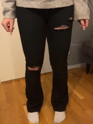 Svarta bootcut jeans med slitningar - Säljer ett par svarta jeans med bootcut-modell och snygga slitningar/hål vid knät och låret. Jeansen har klassisk femficksdesign och är i ett skönt jeans material och sitter jätte bra på Perfekta för dig som gillar en trendig och avslappnad stil.