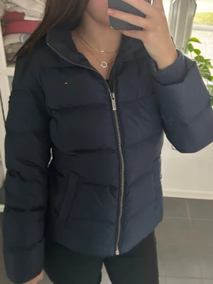 Mörkblå pufferjacka från Tommy Hilfiger - Säljer en mörkblå pufferjacka från Tommy Hilfiger med diskret logga på bröstet och ärmen. Jackan har hög krage, dragkedja framtill och två fickor. Perfekt för kalla dagar då den är gjord på dun. Luvan saknas