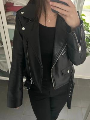 Svart skinnjacka med bälte och dragkedjor - Svart skinnjacka från Pull& Bear med silvriga dragkedjor, bred krage och bälte i midjan. Jackan har flera fickor med dragkedja