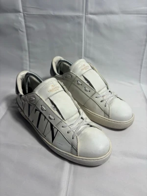 Valentino Garavani skor - Snygga vita Valentino Garavani sneakers i skinn med svart VLTN-logga på sidan och coola nitar runt hälen. Skorna har snörning, rund tå och en silvrig detalj på baksidan. Perfekta för dig som vill sticka ut med exklusiva och trendiga sneakers.