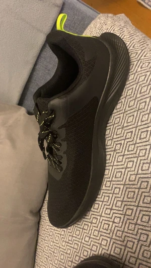 Svarta sneakers med neongula detaljer - Svarta sneakers från Crivit i storlek 46 med neongula detaljer på hälen och snören med gul mönstring. Ovandelen är i mesh och syntet, vilket ger en sportig vibe och bra ventilation. Platt sula och rund tå för skön komfort. Perfekt för dig som gillar stilrena träningsskor.