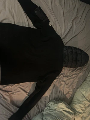 Svart Moncler pufferjacka med huva - Svart pufferjacka från Moncler med stickade ärmar och quiltad front. Jackan har en stor huva, dragkedja framtill och fickor med knappstängning. Moncler-logga på ärmen och extra ficka med dragkedja. Perfekt för dig som vill ha en snygg och varm jacka med street vibe.