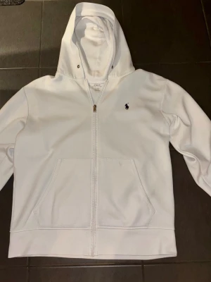 Vit zip-up hoodie från Polo Ralph Lauren, klassisk & stilren - Säljer en stilren vit hoodie med dragkedja från Polo Ralph Lauren. Tröjan har en klassisk passform, huva och kängurufickor. Den ikoniska broderade Polo-loggan finns på bröstet i mörk färg, vilket ger ett snyggt kontrasttryck.