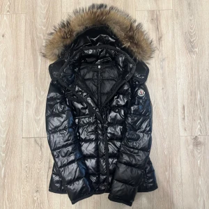 Svart Moncler pufferjacka med päls - Snygg svart pufferjacka från Moncler med glansig finish och fluffig päls på huvan. Jackan har Moncler-logga på ärmen, dragkedja med coola detaljer och knappar framtill. Perfekt för kalla dagar och har en riktigt urban vibe.