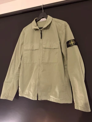 Grön overshirt från Stone Island - Snygg ljusgrön overshirt från Stone Island med klassisk kompasspatch på ärmen. Jackan har två stora bröstfickor, rak passform och dragkedja framtill. Köpt på NK för nypris, väldigt fint skick, storlek L men passar M.