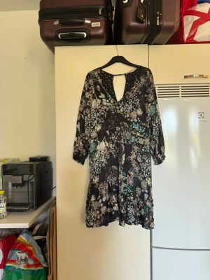 Svart blommig kortklänning med volang - Supersöt svart kortklänning med blommigt mönster i turkos, beige och ljusa toner. Klänningen har v-ringning, långärmade ärmar med resår och volangdetalj längst ner. Materialet känns lätt och flowy, perfekt för dig som gillar boho-vibe och snygga detaljer. Från märket Odd Molly. Storlek 3 som enligt deras storleksguide motsvarar en L. Mycket fint skick, jag hittar inga synliga anmärkningar eller defekter. 