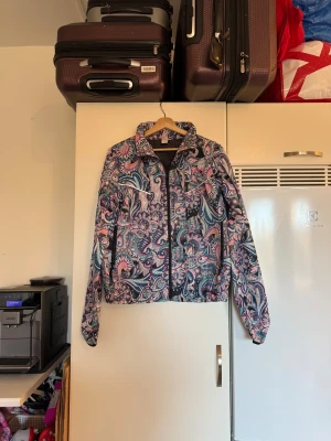 Mönstrad vindjacka från Odd Molly - Säljer en färgstark vindjacka från Odd Molly med unikt blommigt mönster i rosa, blått och lila. Jackan har dragkedja framtill, hög krage och långa ärmar. Perfekt för dig som gillar att sticka ut med coola detaljer och snygg design. Från märket Odd Molly. Storlek 3 som enligt deras storleksguide motsvarar en L. Mycket fint skick, jag hittar inga synliga anmärkningar eller defekter. 