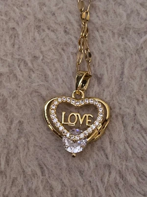 Guldfärgad halsband med hjärthänge - Snyggt halsband i guldfärg med ett hjärthänge dekorerat med glittrande stenar och texten 'LOVE' i mitten. Hänget har en stor klar sten längst ner som ger extra bling. Kedjan är tunn och elegant, perfekt för att lyfta din outfit.