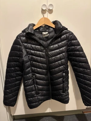 Svart pufferjacka från Mc Kinley - Svart pufferjacka från Everest med huva och dragkedja framtill. Jackan har quiltad design med horisontella och V-formade sömmar, två fickor med dragkedja och reflekterande detaljer baktill. Perfekt för kalla dagar och har en snygg, sportig look.