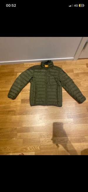 Grön dunjacka Parajumpers YS - Snygg grön dunjacka från Parajumpers i storlek YS. Jackan har quiltad design, hög krage och dragkedja framtill. Klassisk PJS-patch på ärmen och lättviktigt material som håller dig varm. Perfekt för dig som gillar stilren och funktionell look.