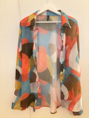 Färgglad mönstrad skjorta PrettyLittleThing - Säljer en transparent och flowy skjorta från PrettyLittleThing i storlek 36. Skjortan har ett abstrakt mönster i blått, rött, gult, brunt och vitt, med klassisk krage och vita knappar framtill och vid ärmslut. Perfekt för dig som gillar färg och statement-plagg eller vill använda som strand skjorta