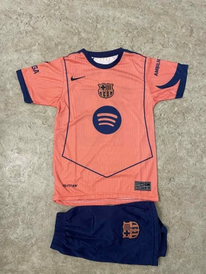 Barça Lamine Yamal fotbollströja set - Säljer ett Barcelona fotbollströja och shorts set med Lamine Yamal #19. Tröjan är korallrosa med marinblå detaljer, klubbmärke och sponsortryck. Shorts är marinblå med klubbmärke. Materialet är lätt polyester, perfekt för fotbollsträning eller match.