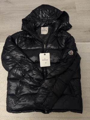 Svart Moncler pufferjacka med huva - Svart pufferjacka från Moncler med glansig finish, huva och dragkedja framtill. Jackan har två sidofickor med dragkedja och Moncler-logga på ärmen. Insidan har en cool serietidningsetikett. Perfekt för dig som vill ha en snygg och varm jacka med street vibe.