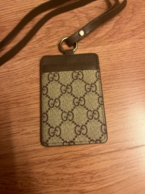  plånbok med GG-mönster - Snygg beige och brun plånbok med klassiskt GG-mönster. Har fönster för ID-kort och en praktisk rem att hänga runt halsen. Tillverkad i skinn och canvas, perfekt för att hålla koll på korten med stil.